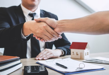 Shaking hands while wearing suits, with documents, calculator and a miniature house on the table