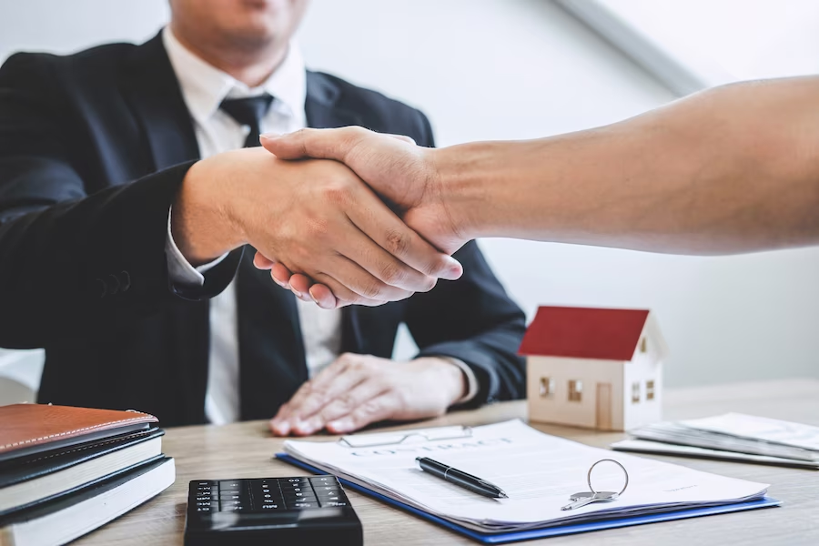 Shaking hands while wearing suits, with documents, calculator and a miniature house on the table