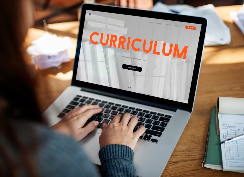 Woman typing on a laptop, screen shows the word "curriculum"