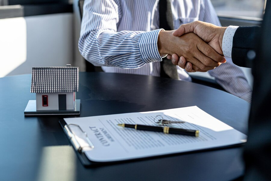 Two individuals shaking hands, with a contract on the table, a pen, and a miniature house